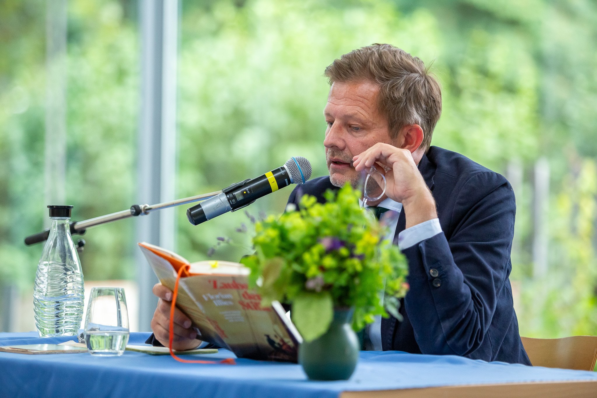 Nahaufnahme von Illies bei der Lesung: Autor mit Mikrofon, Buch, auf dem Tisch vorn Blumen, hinten Wasser.