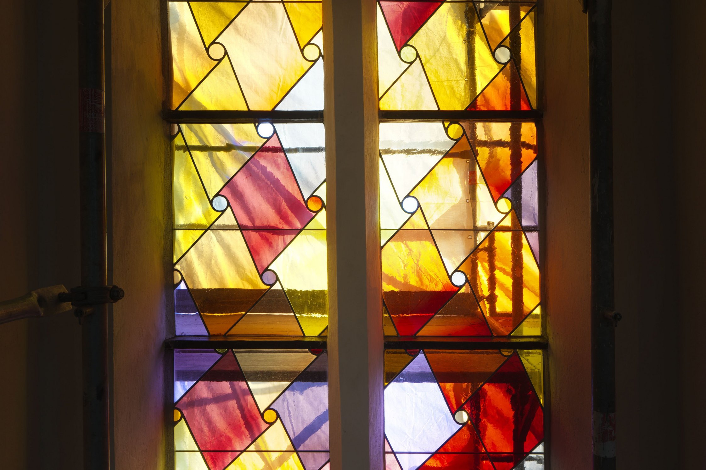 Rot, gelb und blau hinterleuchteter Ausschnitt eiens Kirchenfenster umrandet von einem Baugerüst im Halbdunkel.