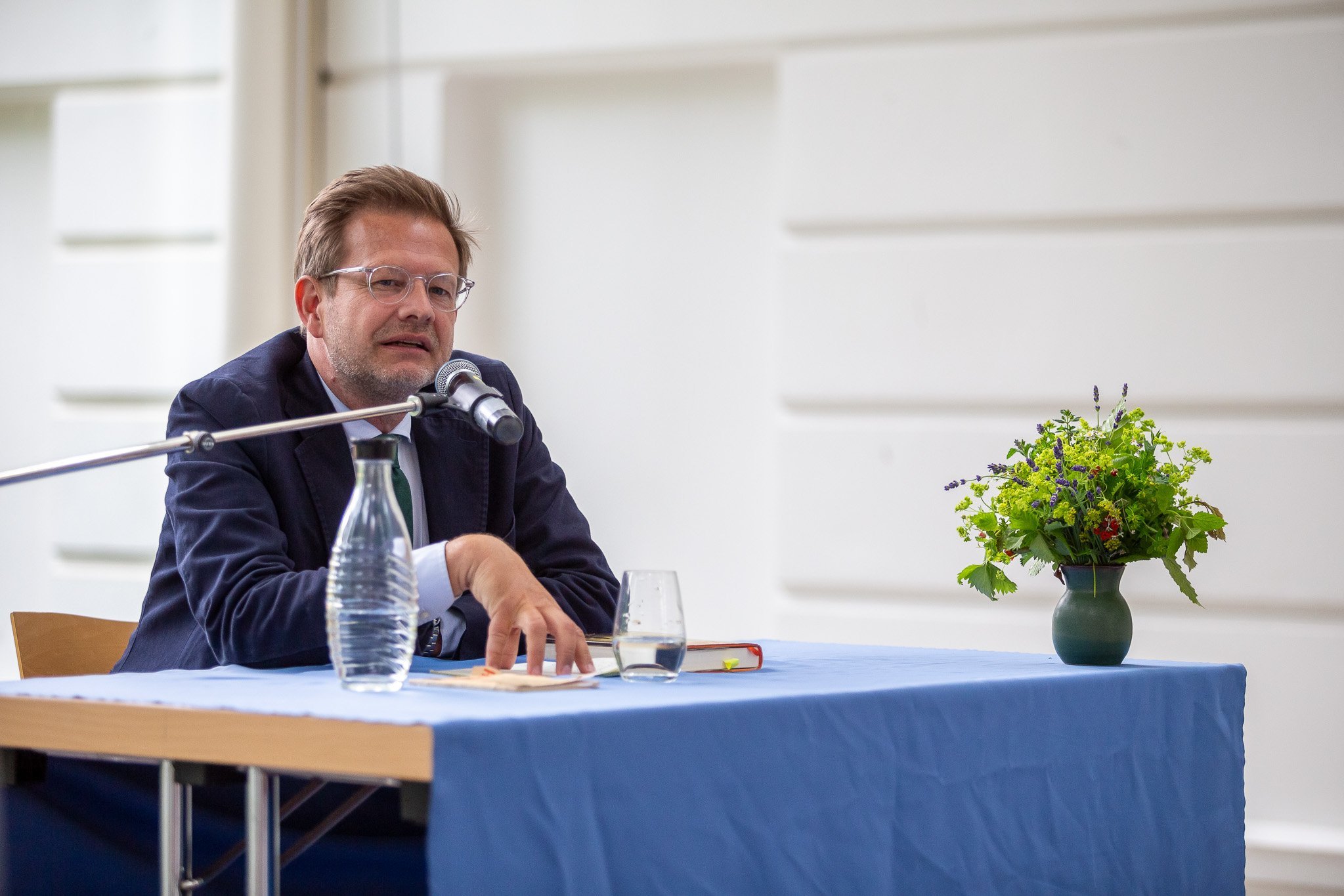 Florian Illies am Tisch: Er spricht und blickt zum Publikum. Auf dem Tisch Wasser, Notizen, Buch und Blumen.